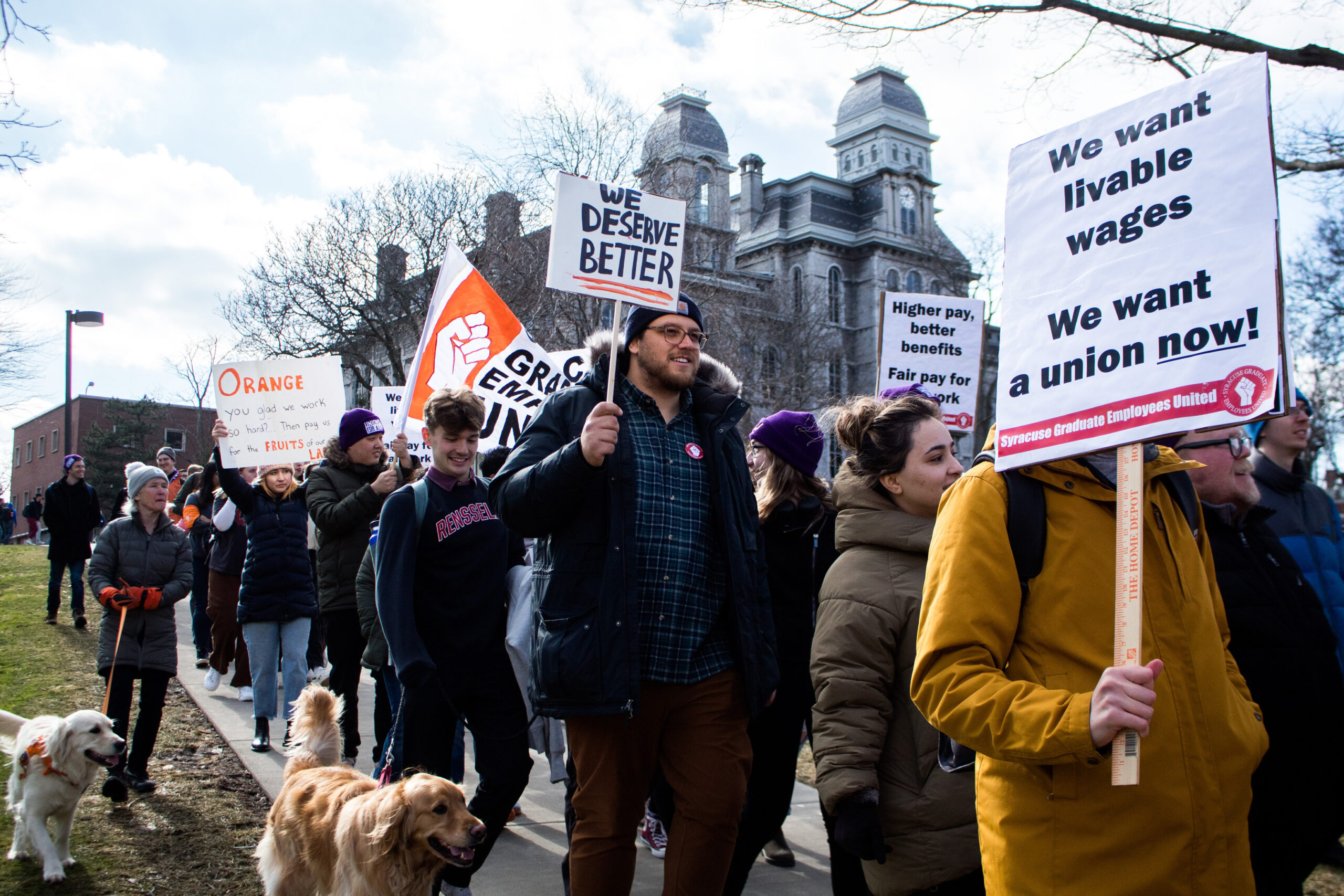 Marchers holding signs and dogs on leashes as they march down a pathway near the Hall of Languages.